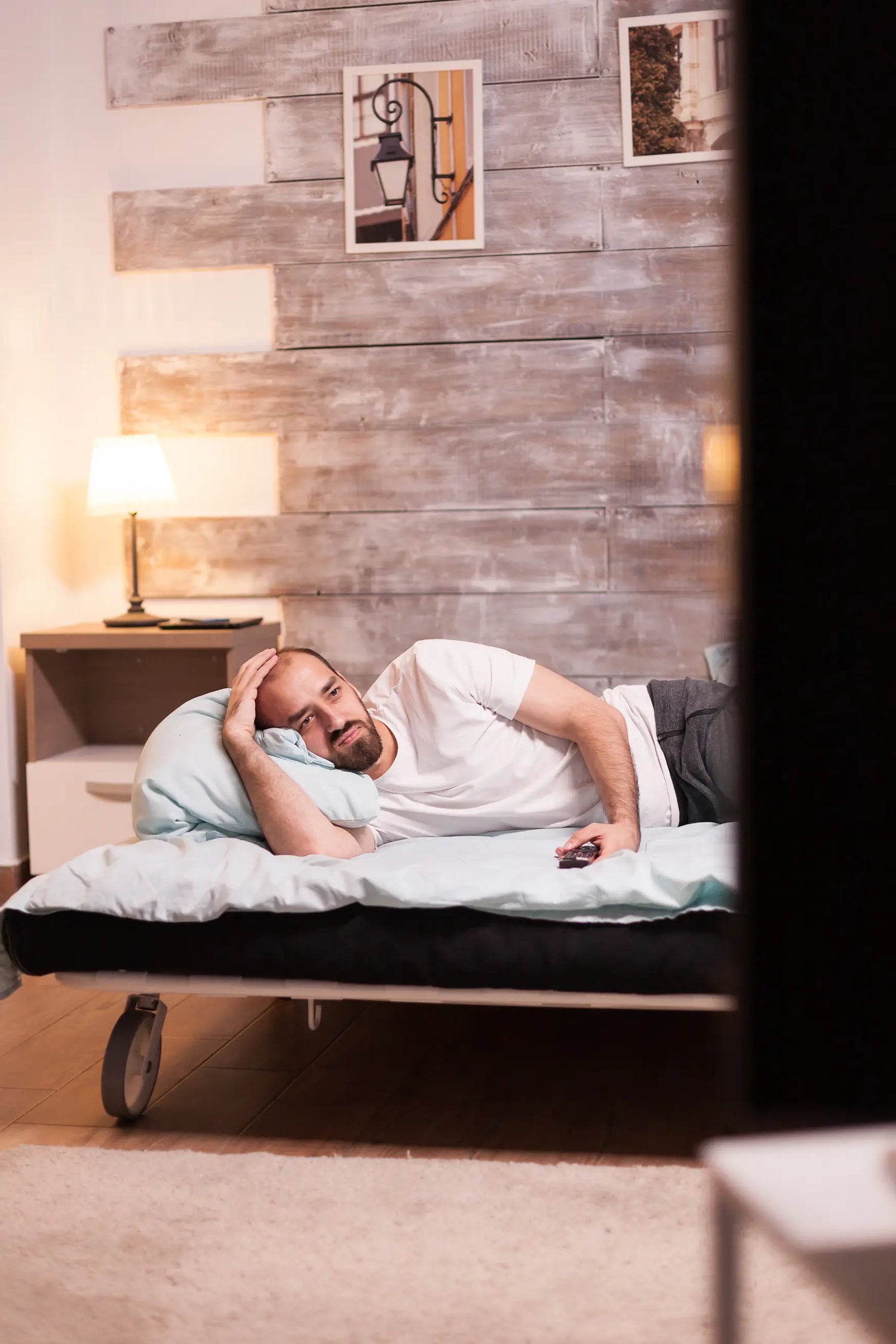 Man suffering from a lack of sleep lying on a bed in a room with wooden walls and a lamp.