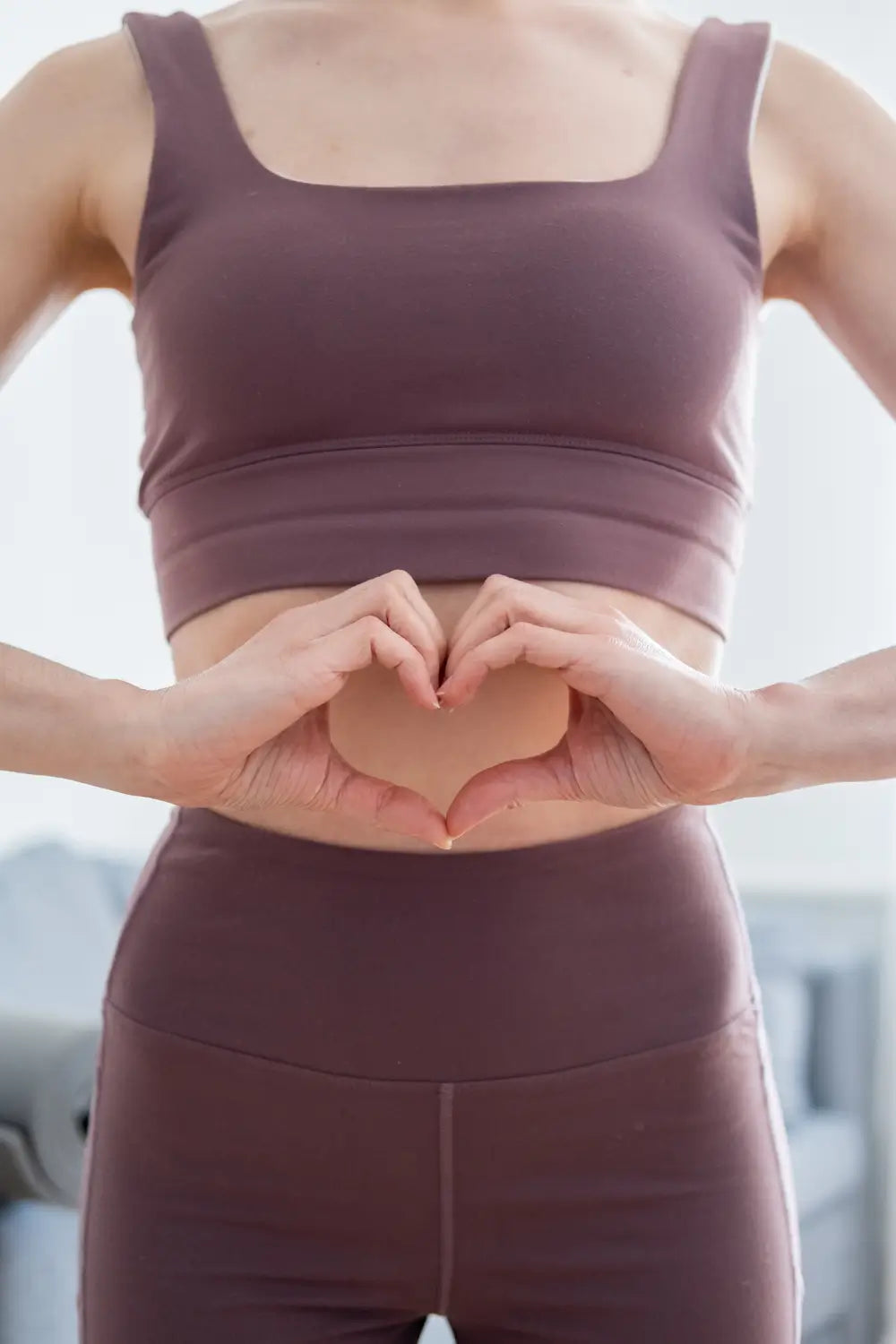 Woman wearing a brown athletic outfit making a heart shape in front of her stomach with herhands.