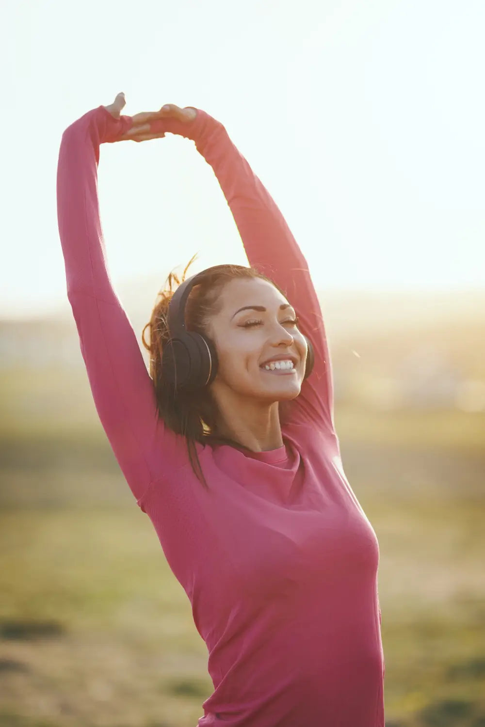 Woman in pink shirt with headphones, stretching outdoors with a blurred natural background